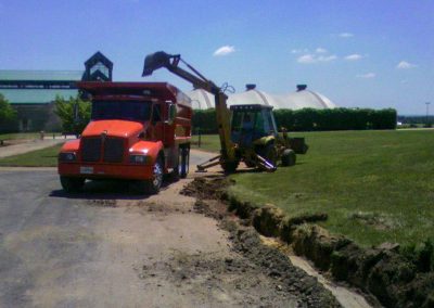 A backhoe from an asphalt paving company is digging a trench next to a red dump truck on a sunny day, with buildings and a green lawn in the background.