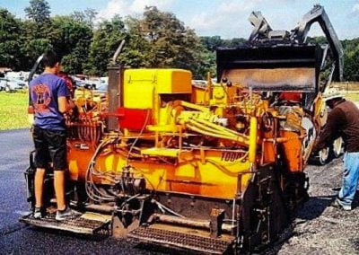 Workers from a reputable paving contractor are using a yellow asphalt paving machine on a sunny day in Frederick, Maryland, with trees and a blue sky in the background.