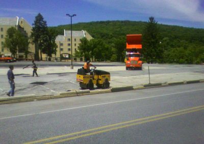 Construction workers perform asphalt paving on a parking lot while an orange truck with its hood open stands in the background, capturing a typical scene in Maryland.
