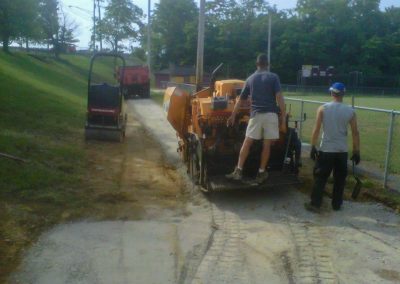 Two workers from a Maryland paving contractor operate machinery to pave a narrow road by a grassy area and a baseball field. A red truck is in the background.
