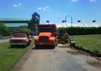 A backhoe digs along a paved path next to two orange trucks from an asphalt paving company near large greenhouses and buildings on a sunny day.
