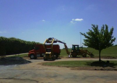 A backhoe loader loads dirt into a dump truck at a construction site in Frederick, MD, under a clear blue sky. Nearby, workers prepare for asphalt paving.