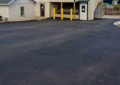 Two-story building with a green roof, tan siding, and yellow bollards near an entry door. The driveway was recently completed by an MD asphalt paving company.