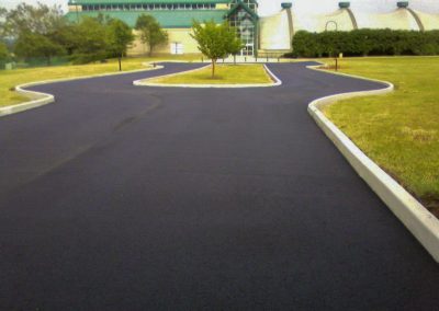 Newly paved, winding asphalt road bordered by fresh grass, leading to a green-roofed building and trees in the distance, showcasing the exceptional work of an asphalt paving company in Frederick, MD.