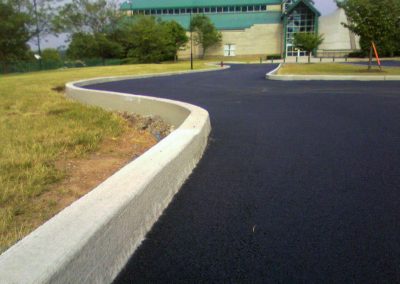 A freshly paved, winding road leads to a modern building with a green roof, surrounded by trees and grass, thanks to an expert MD asphalt paving company.
