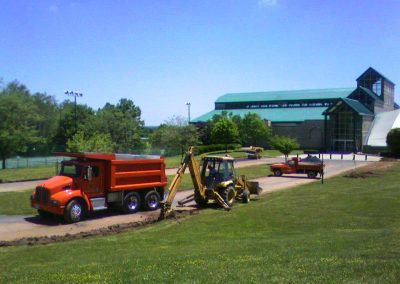 The construction site in Frederick, Maryland, features a red dump truck and an excavator digging a trench near a large building with a green roof, preparing the area for asphalt paving.