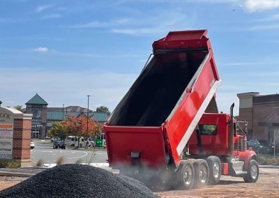 A red dump truck unloading gravel onto a construction site with buildings in the background under a blue sky, prepared for an asphalt paving company in Maryland.