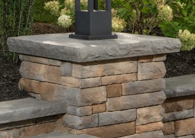 Stone pillar with a black lantern on top, surrounded by greenery and flowers in a landscaped garden setting in Frederick, Maryland. The beautiful arrangement was expertly crafted by a local paving contractor.