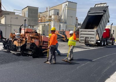 Workers in safety gear from an asphalt paving company lay asphalt on a road using heavy machinery and tools, with buildings in the background, in Frederick, MD.