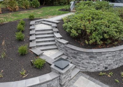 A stone pathway and steps, complemented by landscaped shrubs and a retaining wall, create an inviting entrance in this residential garden. For durability and aesthetic appeal, consider an asphalt paving company for the driveway's finish.