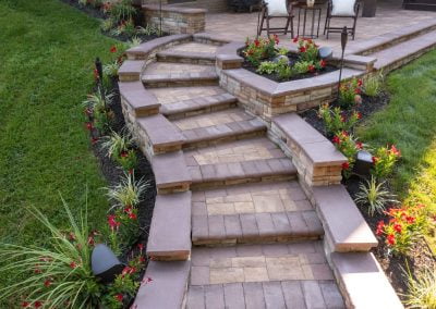Curved stone steps with red flowers and green plants on either side lead up to a patio with chairs and a table, meticulously designed by a local Maryland paving contractor.