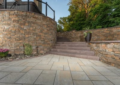Outdoor stone patio with stairs leading to an upper level and potted plants. Trees and greenery in the background add a touch of nature, while the adjacent asphalt paving provides a smooth transition to other areas. Consider hiring an asphalt paving company in MD for professional results.