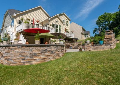 A beautiful backyard in Frederick, MD, with a patio, stone retaining wall, umbrellas, plants, and a large two-story house.