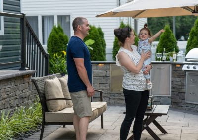 A smiling family in a backyard: a man, a woman holding a toddler who is pointing, with outdoor furniture and a grill nearby. Their pristine patio was crafted by an expert asphalt paving company in MD.