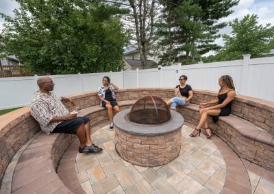 Four people sitting and chatting around a round, brick fire pit in a backyard with greenery and a white fence in Frederick, MD.