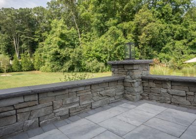 A stone patio with a low stone wall and a lantern, overlooking a lush green garden with tall trees in the background, crafted by a skilled Frederick paving contractor.