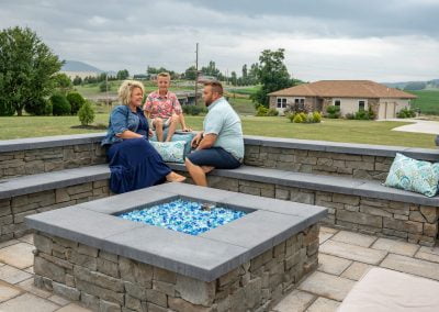 Three people sitting around a stone fire pit in a Maryland backyard with green lawn and cloudy sky in the background.