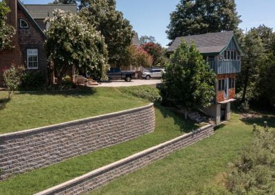 Two houses next to each other in Frederick, separated by a tiered retaining wall crafted by a skilled paving contractor, with a grassy slope and trees in the background.