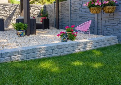 Outdoor patio with a stone wall, potted pink flowers, wicker furniture, pink chair, and hanging flower baskets, all beautifully complemented by the smooth finish provided by an expert asphalt paving company.