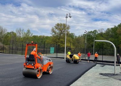 Construction workers in orange vests from a Maryland asphalt paving company use heavy machinery to pave a road on a cloudy day. Trees and fences are in the background.