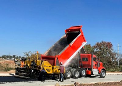 A red dump truck pours materials into a yellow paver on a sunny day as workers stand nearby on a Frederick construction site managed by a skilled paving contractor in Maryland.