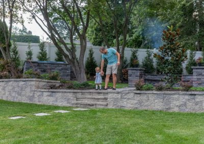 An adult helps a child step down stone steps in a landscaped backyard with greenery and trees, creating a serene and picturesque setting reminiscent of the charming estates found in Frederick, MD.