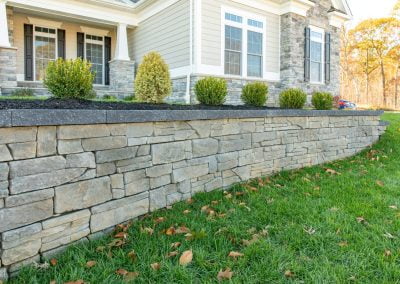 Stone retaining wall with shrubs in front of a house featuring stone accents and beige siding, all beautifully framed by green grass. Crafted by an expert paving contractor in MD, the landscape showcases meticulous attention to detail and quality.