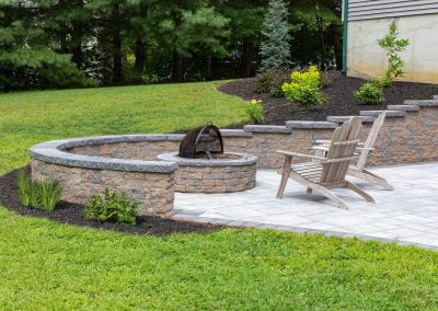 Stone patio with a fire pit, wooden Adirondack chairs, and landscaped garden beds in a lush green backyard setting, crafted with expertise by an MD paving contractor.