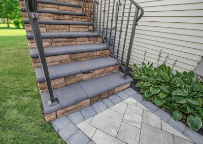 Outdoor stone stairs with black metal railings lead up to a house with siding. Green plants grow beside the stairs, showcasing the meticulous work of an MD paving contractor.