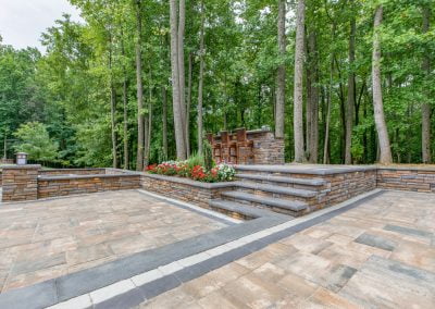 Outdoor patio with stone steps leading to a bar area surrounded by tall trees and flower beds, showcasing high-quality work reminiscent of a top Maryland asphalt paving company.