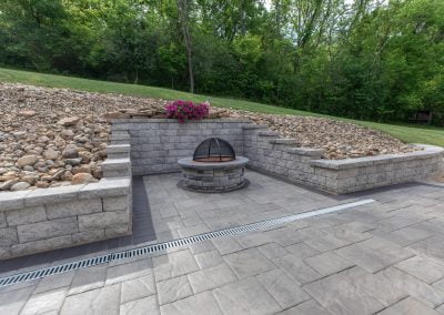 Stone patio with a built-in fire pit, surrounded by a retaining wall with decorative rocks and a flower pot. Crafted by an expert paving contractor in Maryland (MD).