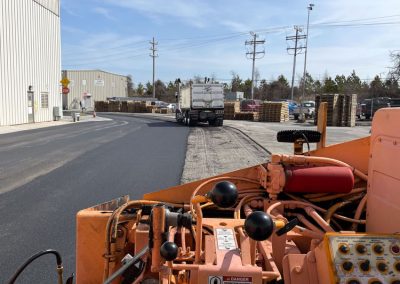 View from a paving machine operator's seat, showing freshly paved asphalt and a truck, with buildings under a clear Maryland sky.