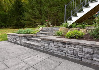 In Frederick, Maryland, a backyard with stone steps and a retaining wall surrounded by greenery leads to a white staircase with black railing. An asphalt paving company added complementary pathways, enhancing the outdoor space's charm.