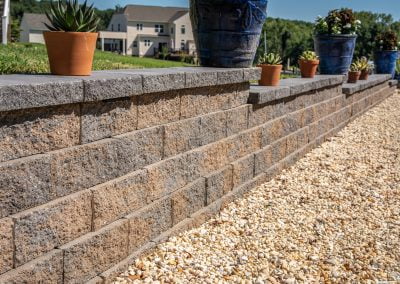 A stone retaining wall with potted plants on top and a gravel pathway beside it, crafted by a paving contractor in Maryland, with houses and trees in the background.