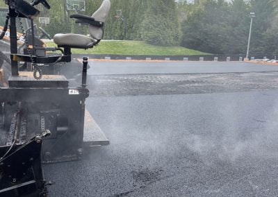 A paving contractor's machine is laying fresh asphalt on a roadway next to a grassy area, with basketball courts in the background.