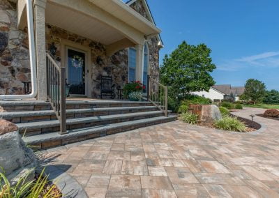 Stone porch with stairs, a black chair, and potted plants on a sunny Maryland day, leading to a landscaped front yard.