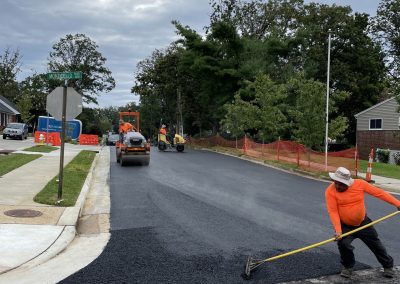 Workers repaving a residential street; one smooths asphalt with a rake while machinery operates behind.
