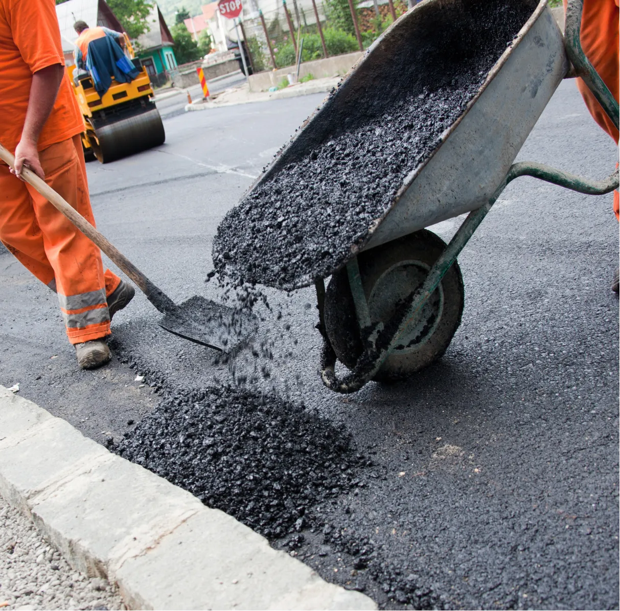 asphalt-millings-3 Workers in orange uniforms paving a road with asphalt using a wheelbarrow and shovel.