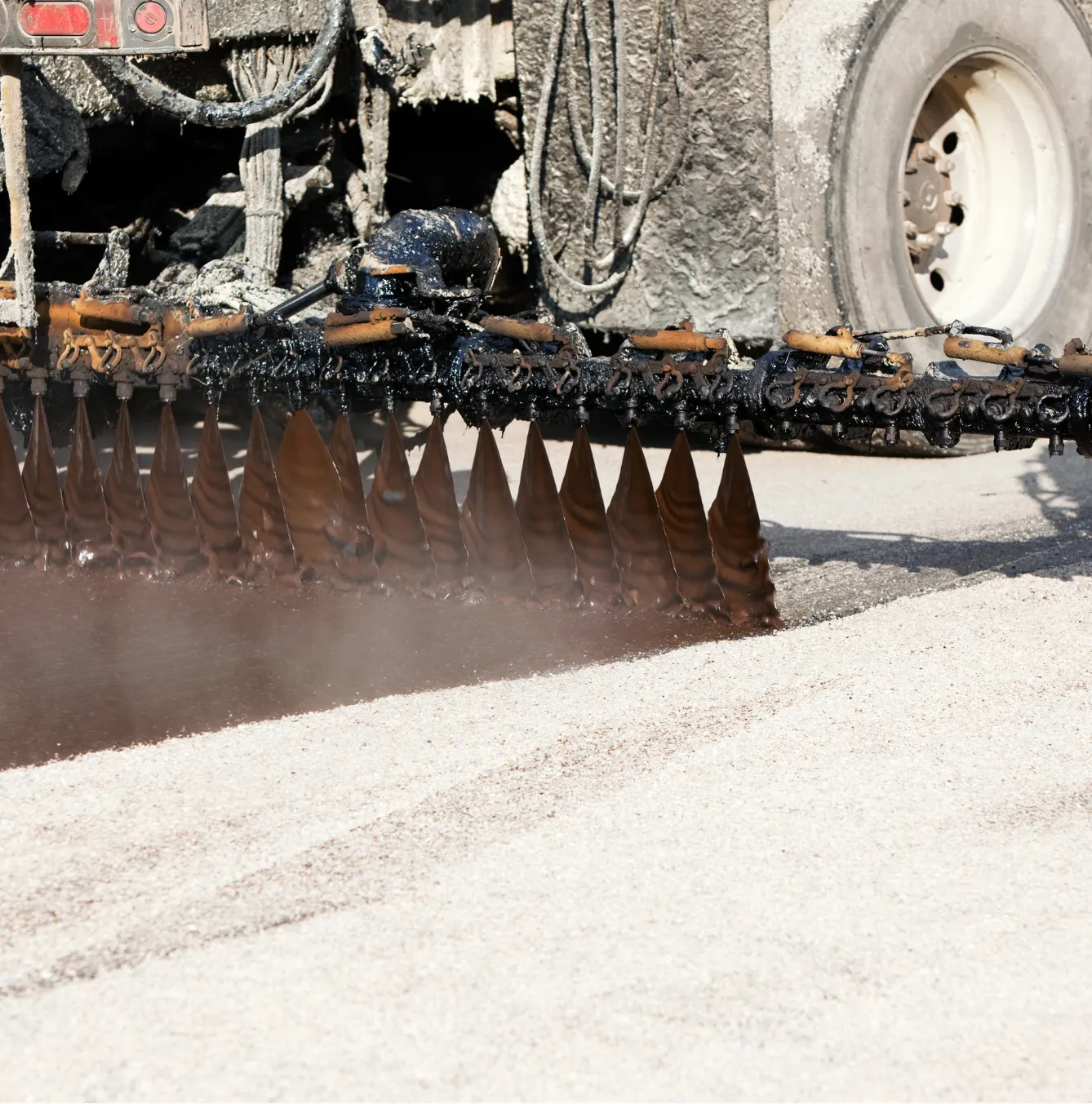 tar-and-chip-1 Machine spraying tar on a road during construction with visible wheels and hoses.