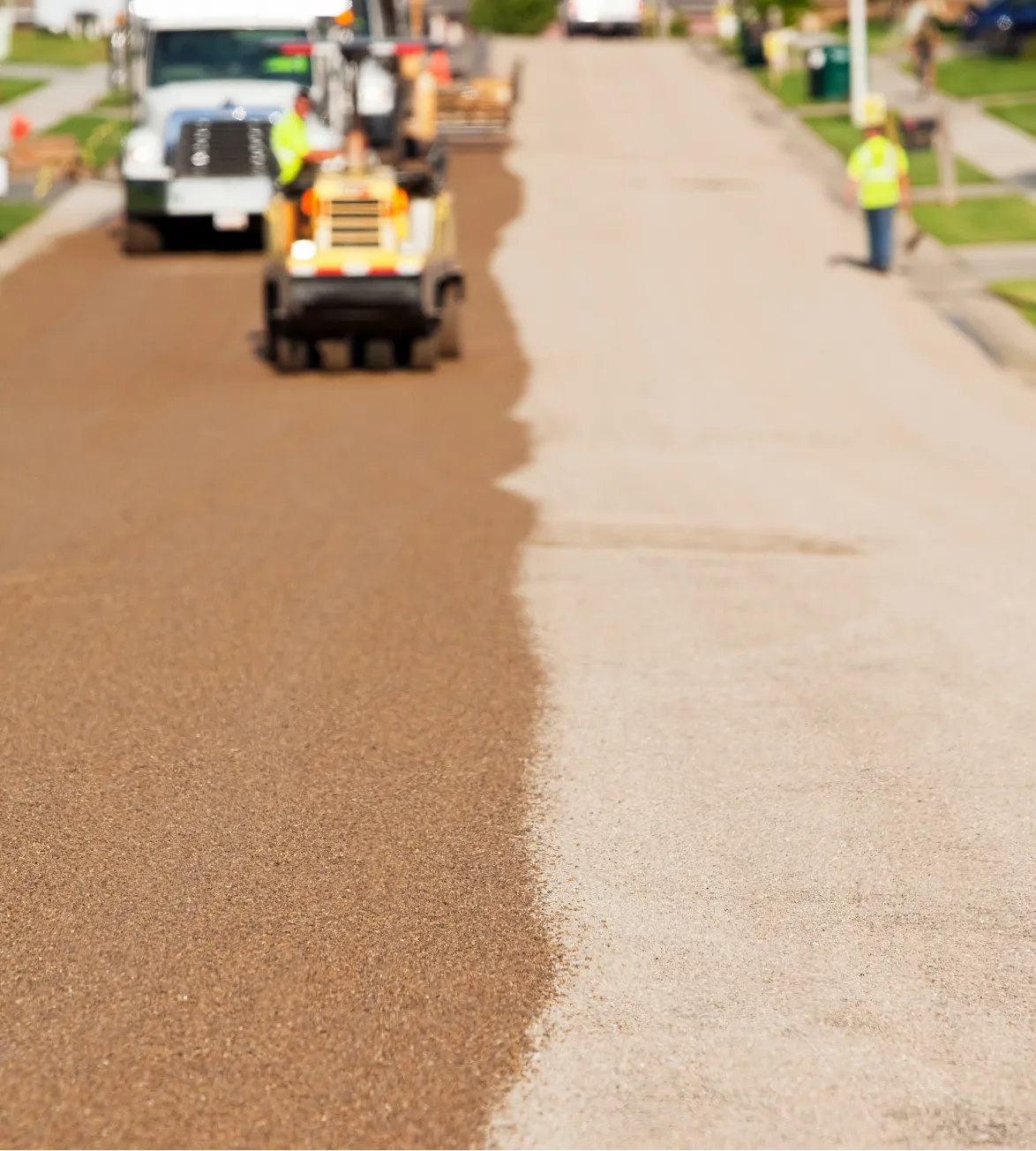 tar-and-chip-2 Construction workers paving a residential street with machinery; half paved and half unpaved surface visible.