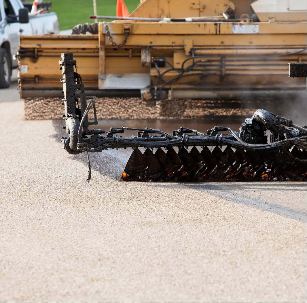 tar-and-chip-3 Close-up of a paving machine laying fresh asphalt on a road, with visible machinery detail and smoke.
