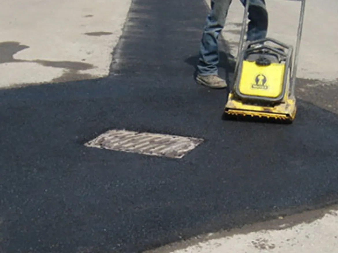 maintenance-1 A person uses a compactor to smooth fresh asphalt around a manhole cover on a road.