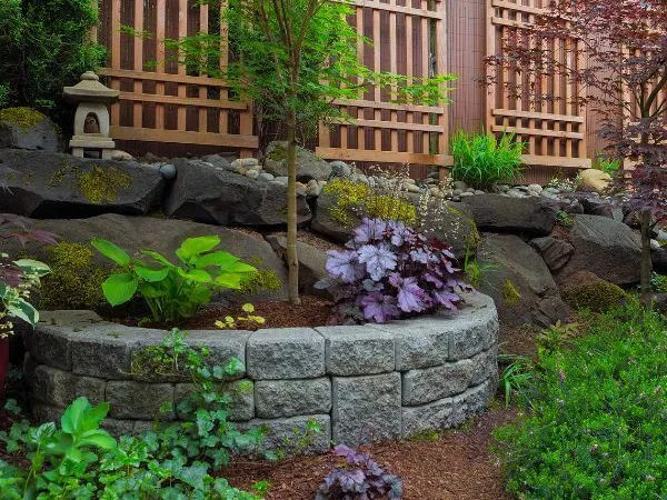 hardscape-1 A stone-bordered garden bed with green and purple plants, set against a wooden fence and rocks.
