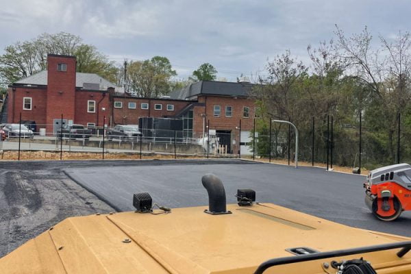 A construction vehicle paves fresh asphalt near a red brick building on a cloudy day.