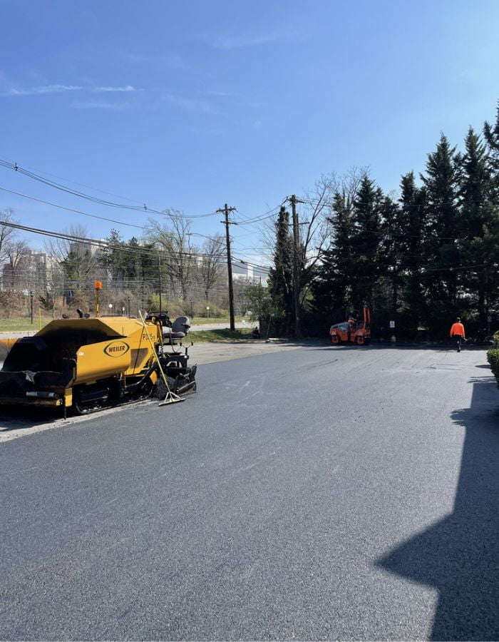 Copy-of-ceemitemplate-700-900 Fresh asphalt being laid on a road with paving equipment and workers under a clear blue sky.