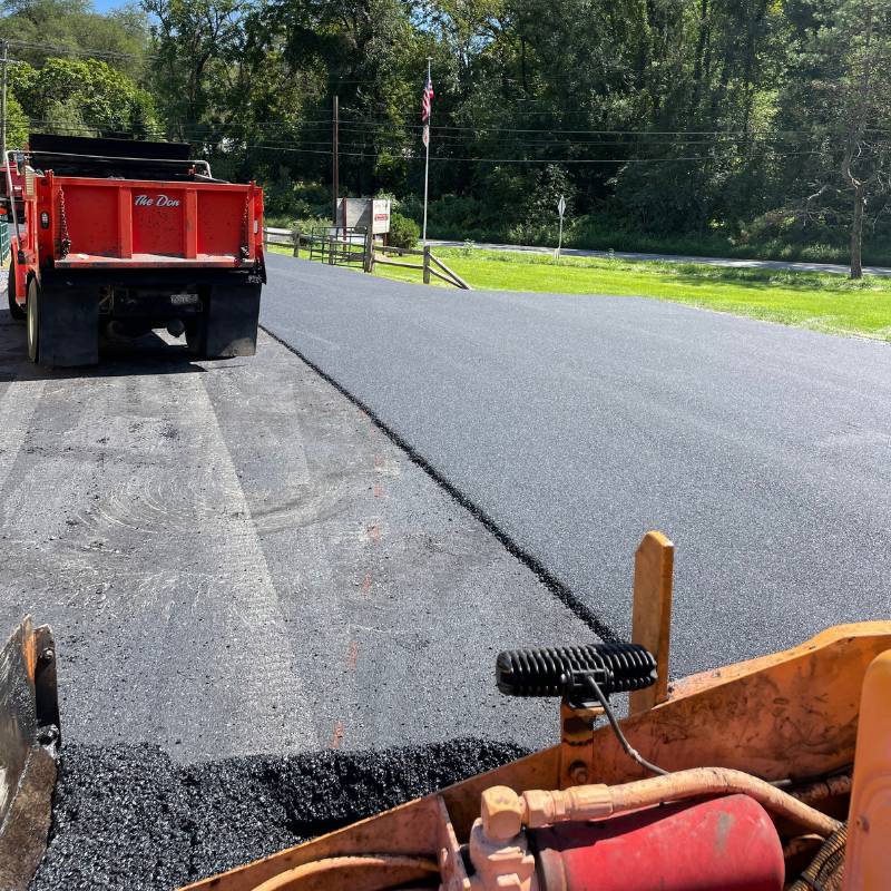 xr:d:DAFj4qgEvJY:29,j:5419394044,t:23052503 Crews repaving a road with fresh asphalt on a sunny day; a dump truck and paving equipment visible.