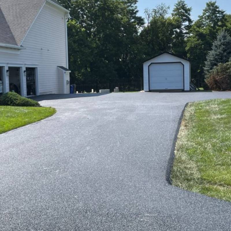 xr:d:DAFj4qgEvJY:30,j:5419409891,t:23052503 Freshly paved driveway leading to a white garage and a small shed, bordered by green grass and trees.