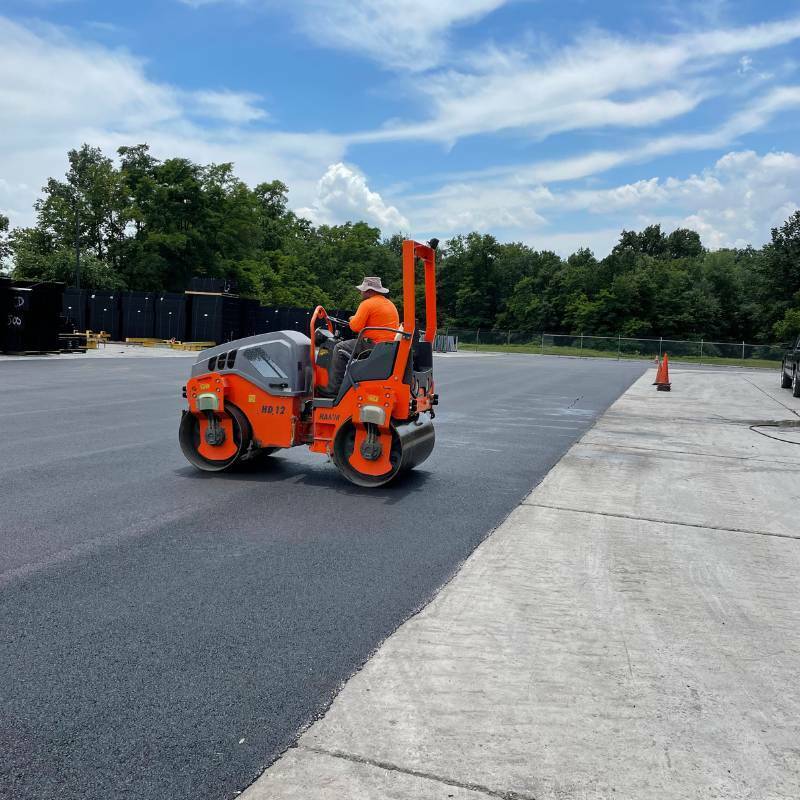 A worker operates an orange steamroller to pave fresh asphalt under a partly cloudy sky.
