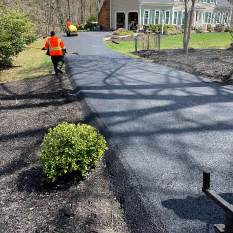 Two workers paving a driveway with fresh asphalt near a house surrounded by greenery.