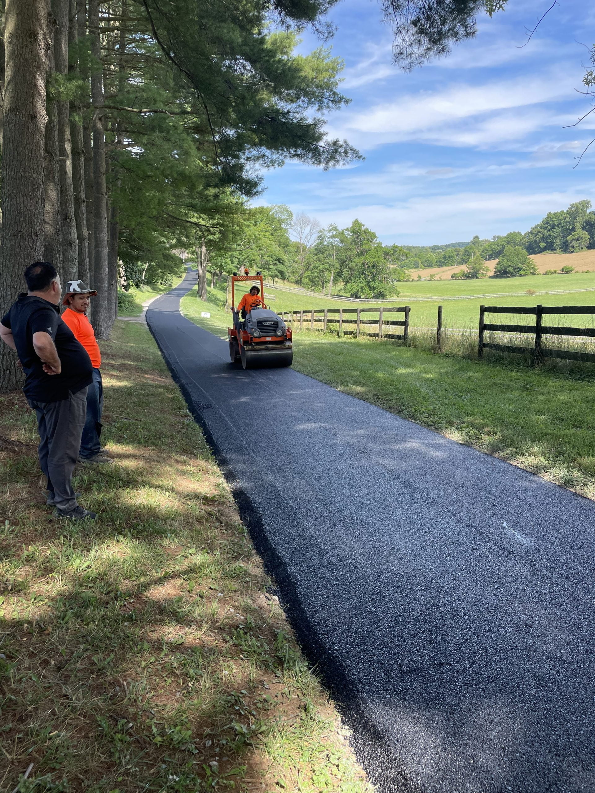 IMG_4285-1-scaled-1 Two workers watch as a person operates a roller on a newly paved rural road lined with trees and fields.
